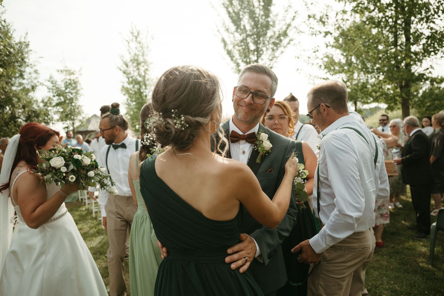 Guests mingling outdoors at a wedding reception, with a smiling groom in a green suit and bow tie greeting attendees.