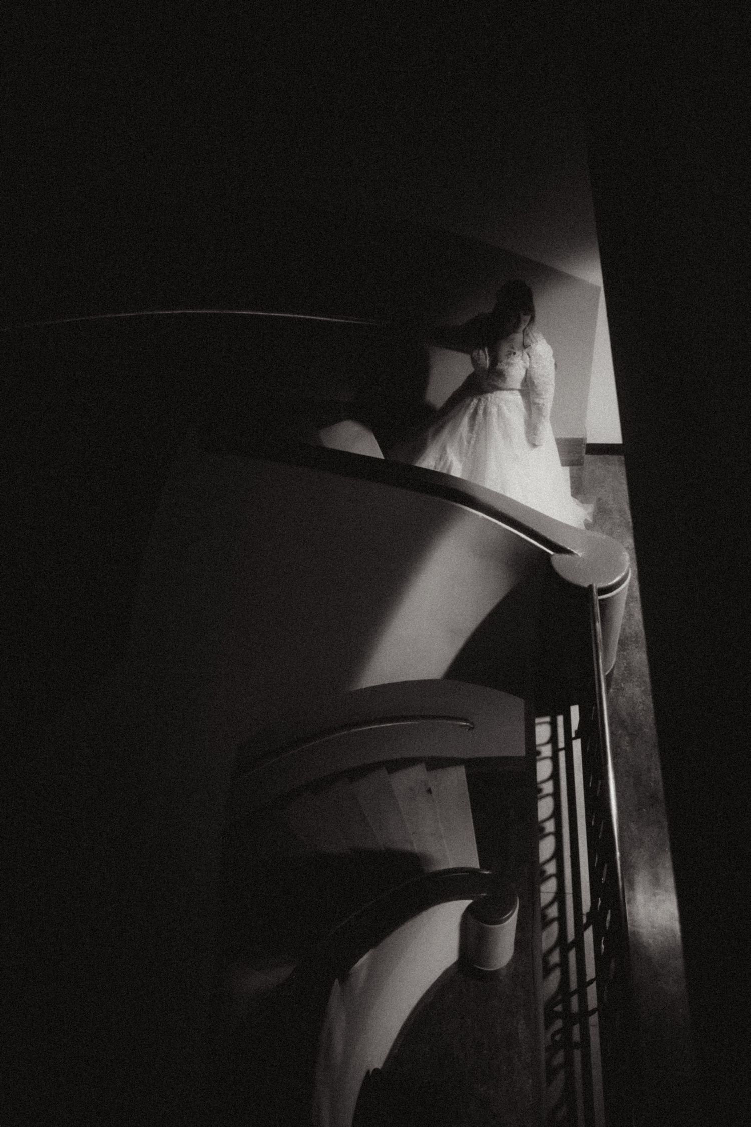 Bride in white dress ascending a dramatic spiral staircase in moody black and white wedding photography.
