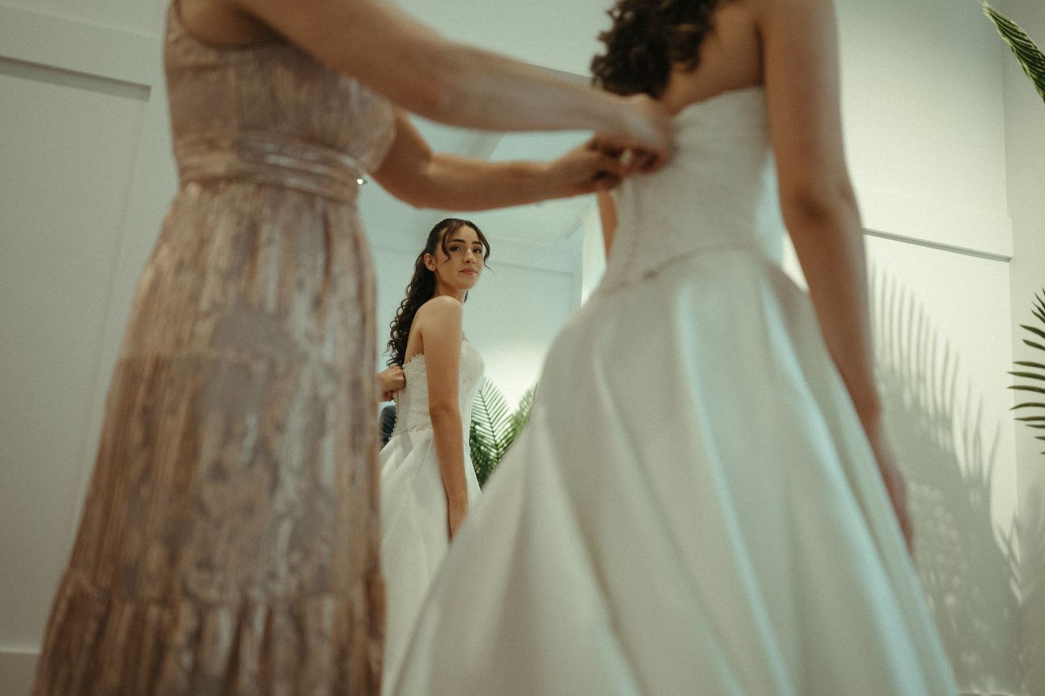 Bridesmaid helping bride into white wedding gown reflected in mirror during bridal preparations.