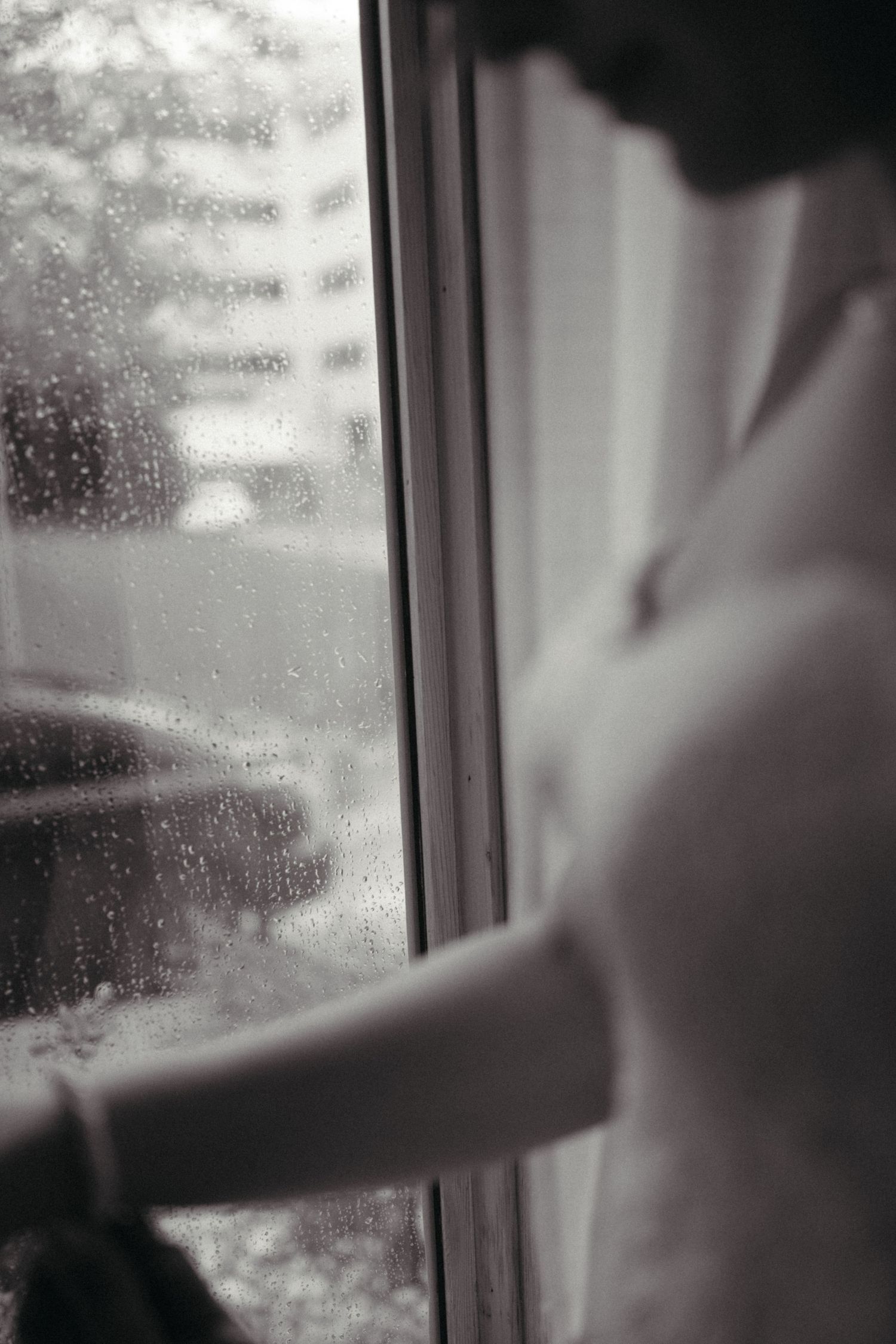 Black and white photo of a bride standing by a rain-covered window in her wedding dress.