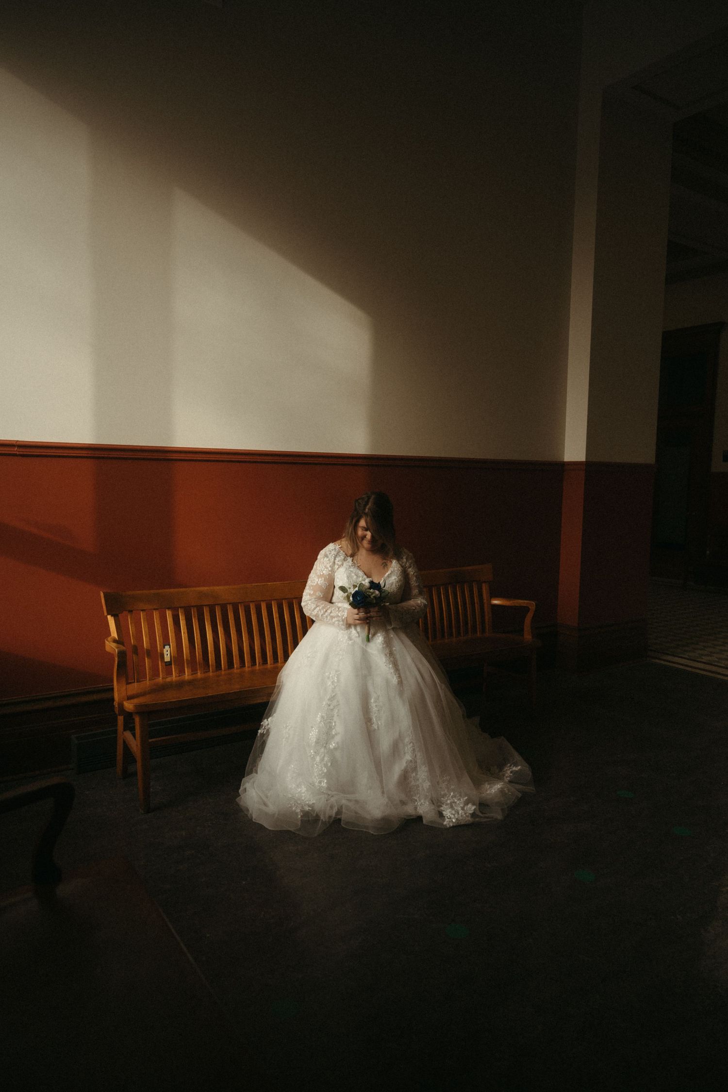 Bride in lace ball gown holding bouquet standing in dramatic light near wooden benches in elegant venue.