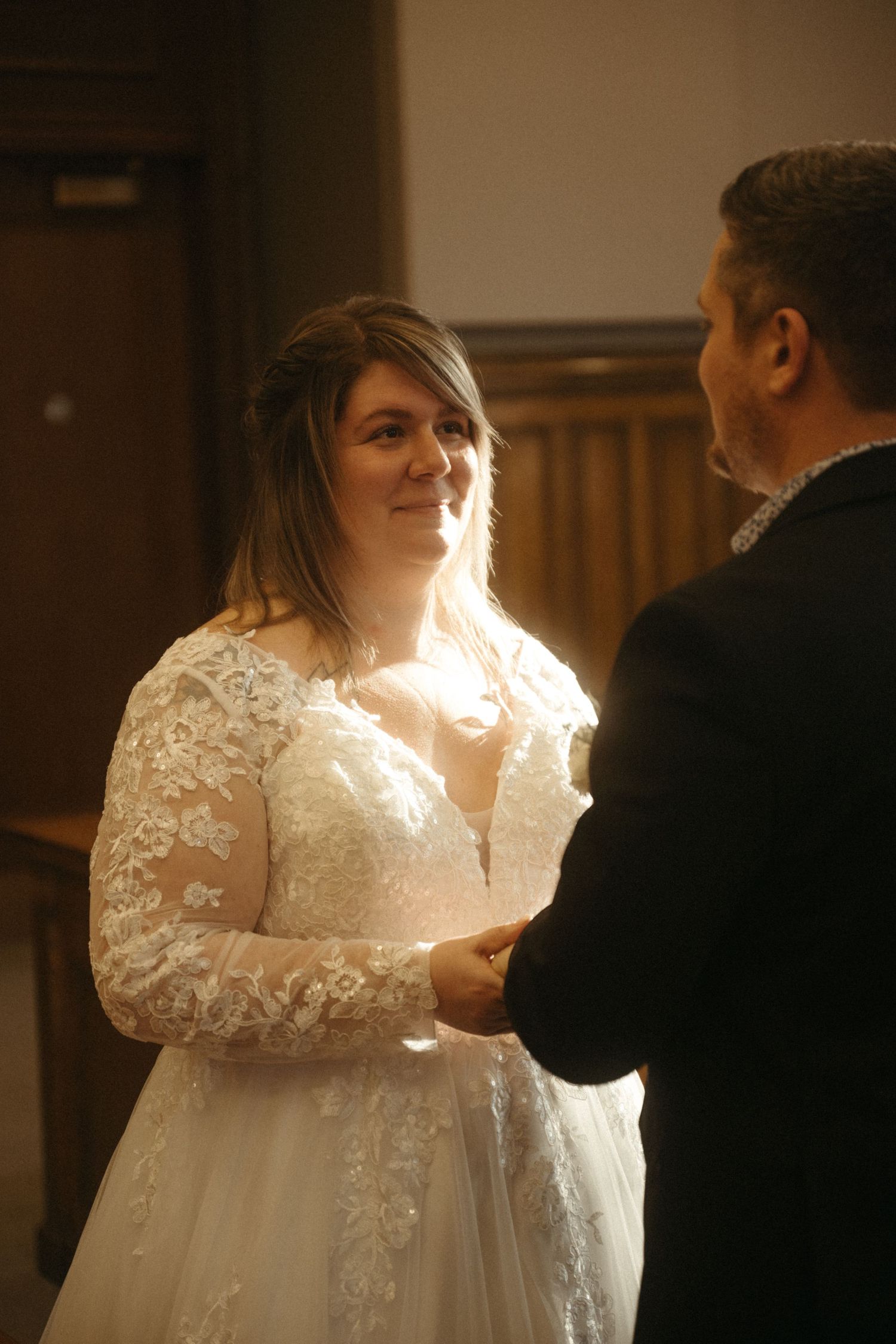 Bride in lace wedding gown smiling at groom during indoor wedding ceremony.