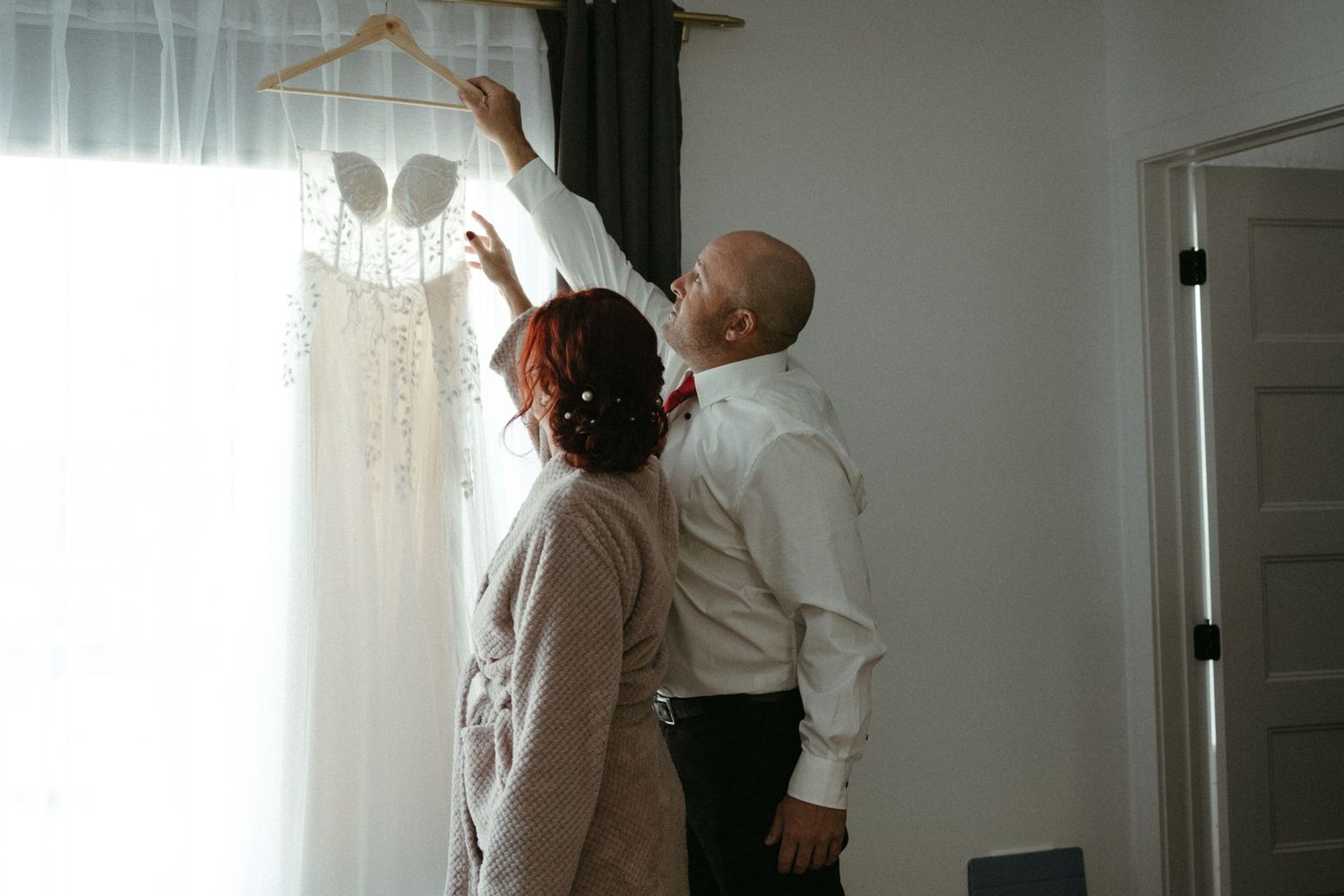 Couple hanging a wedding dress on a hanger near a bright window in a hotel room.