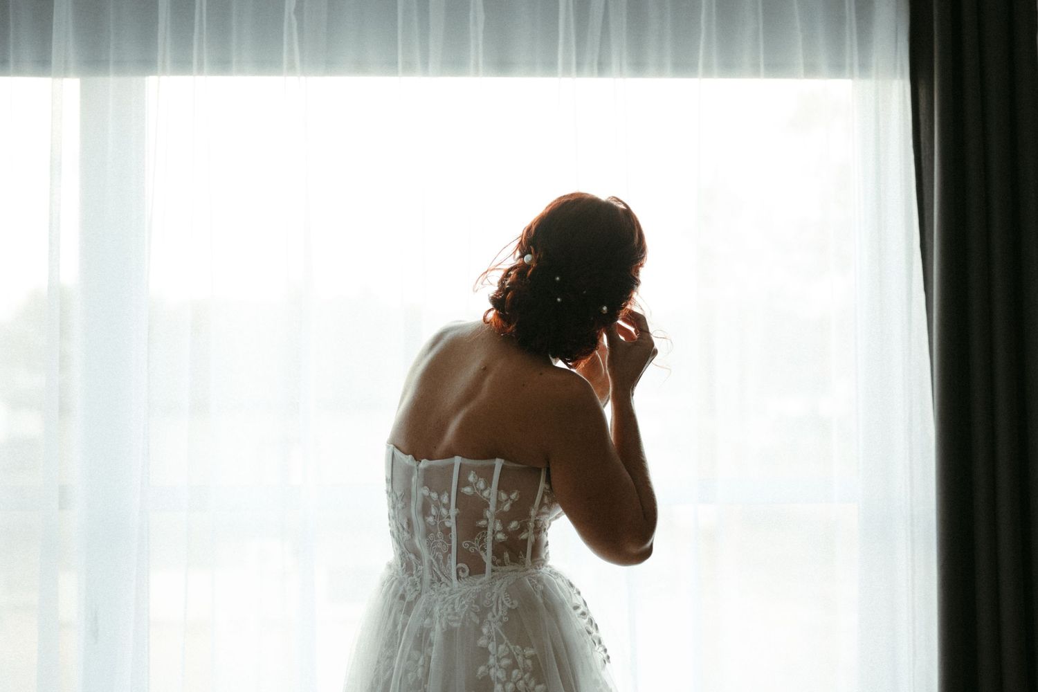 Bride in lace corset wedding gown stands by sheer curtained window adjusting her earring.