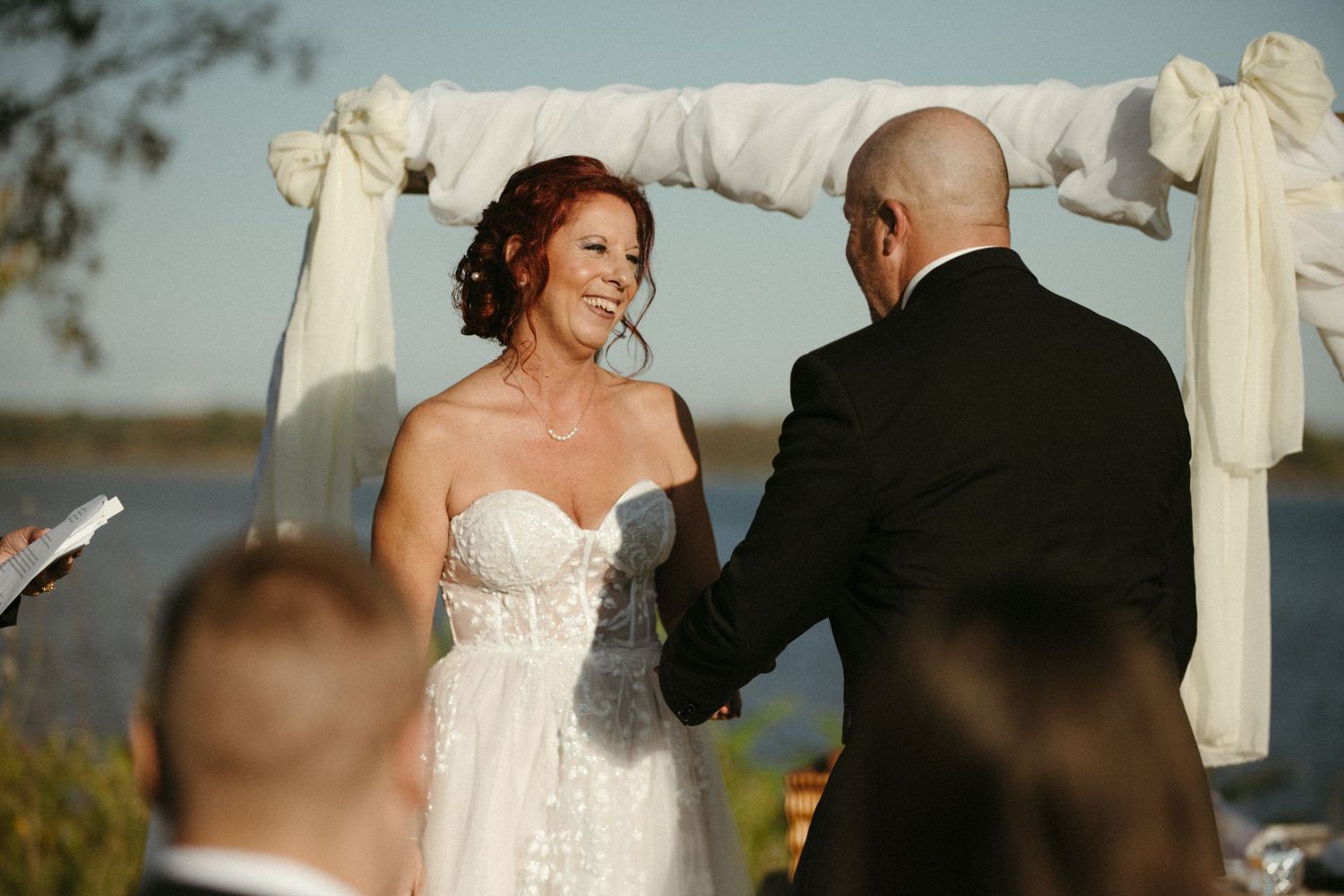 Bride in strapless lace gown holds hands with groom at outdoor wedding ceremony under white draped arch.