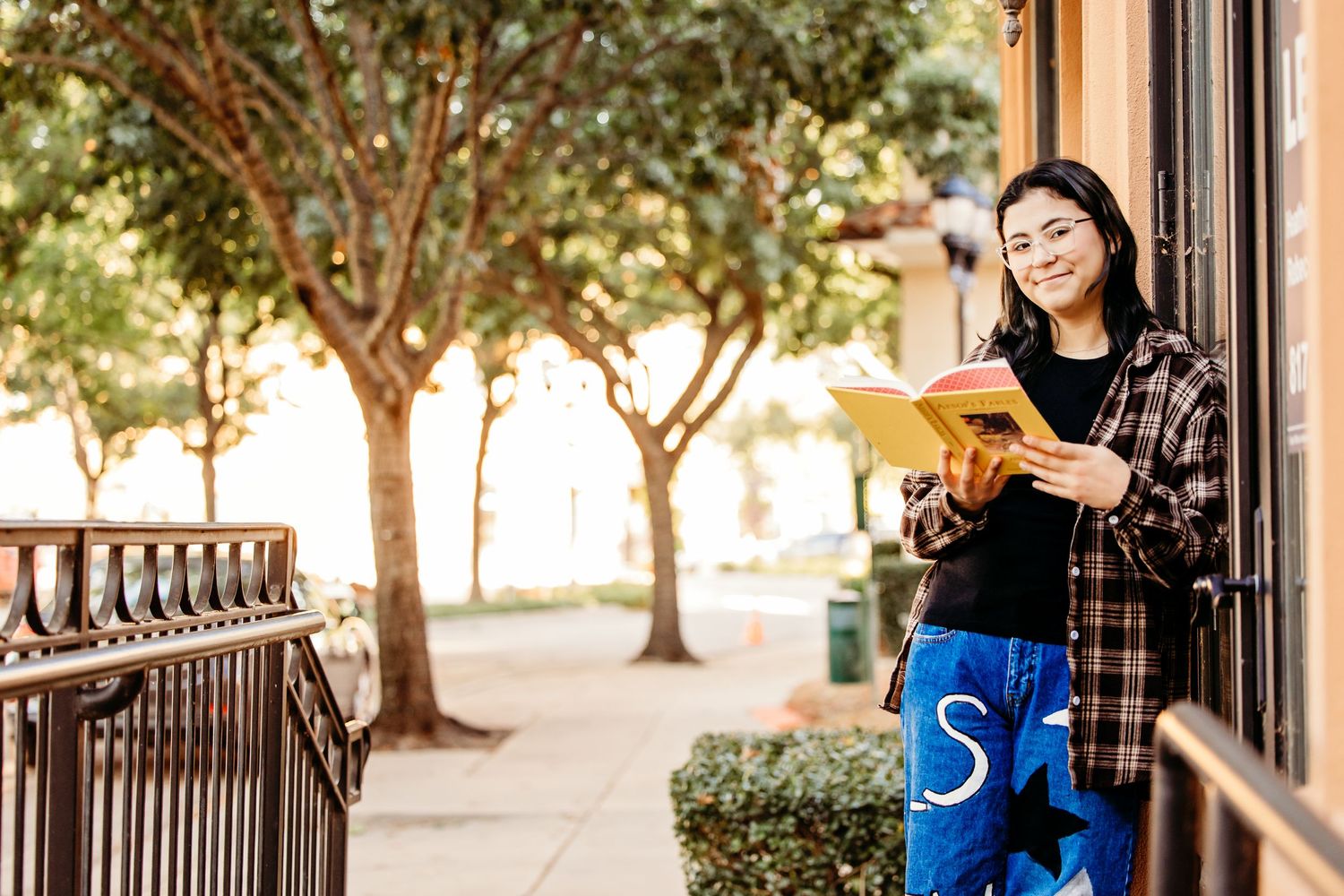 Senior girl leans against the side of a quaint shoppe in Colleyville while holding an open book.