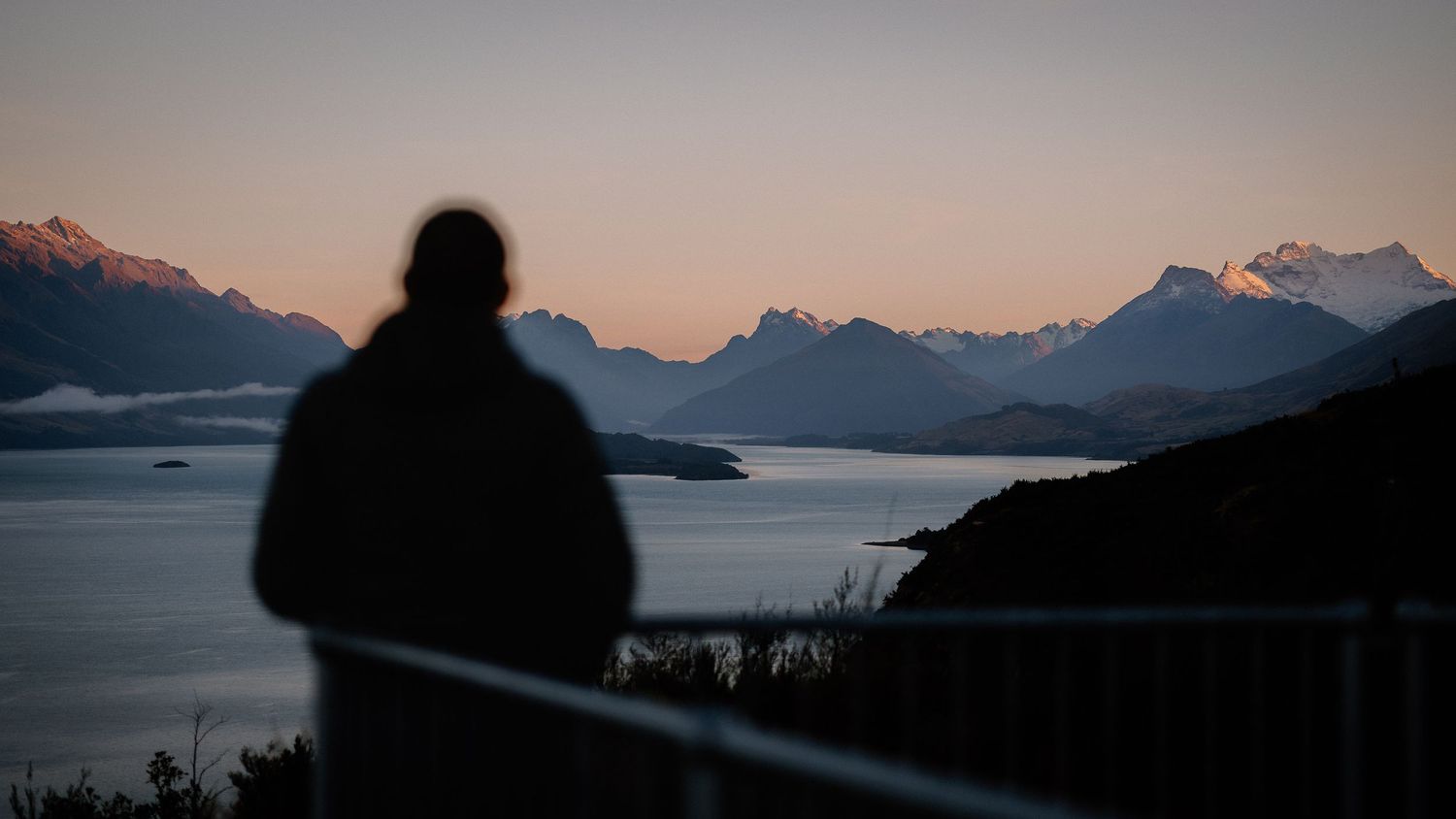 Mountain peaks silhouetted against a colorful sunset sky reflect in calm lake waters at dusk.