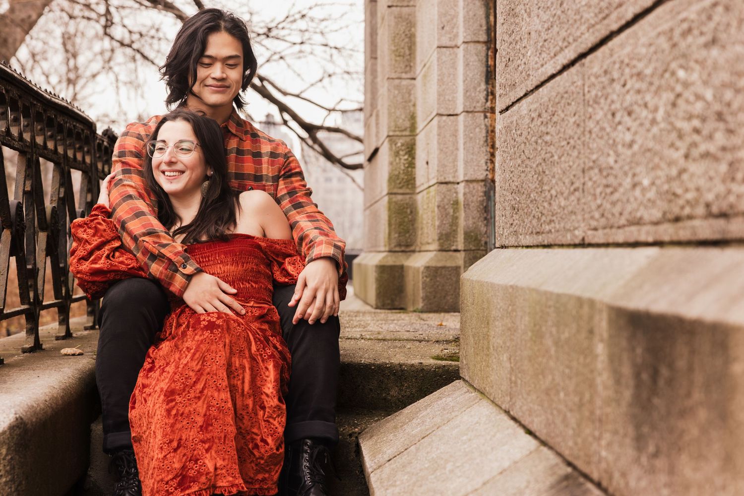 A couple in coordinating orange and plaid outfits pose together against an urban skyline backdrop for engagement photos.