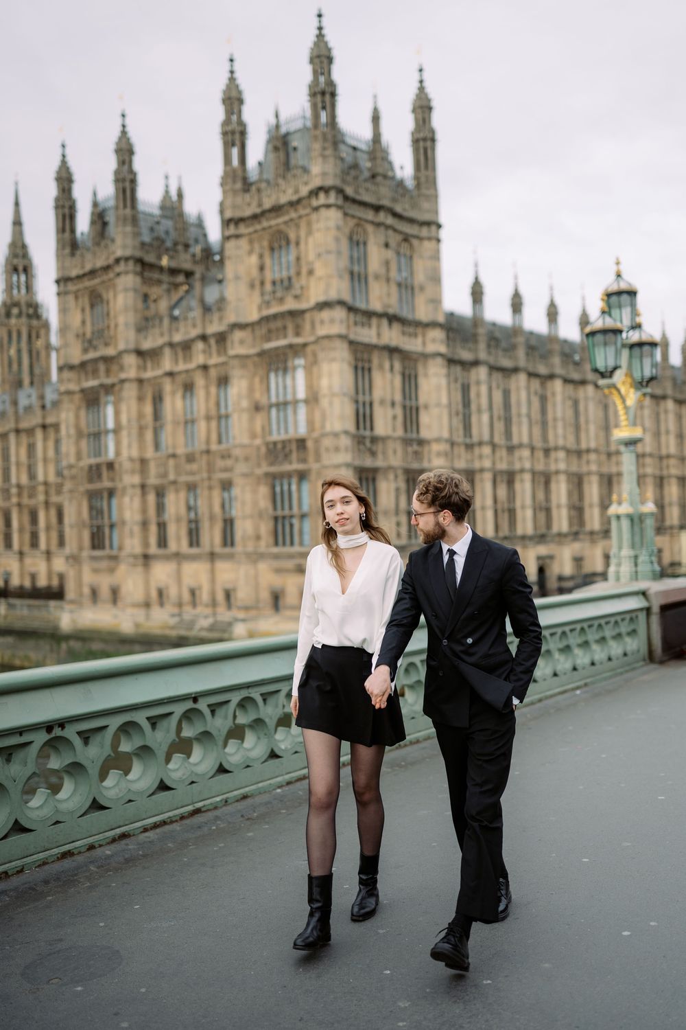 A couple walks together on a bridge near the Houses of Parliament in London on an overcast day.