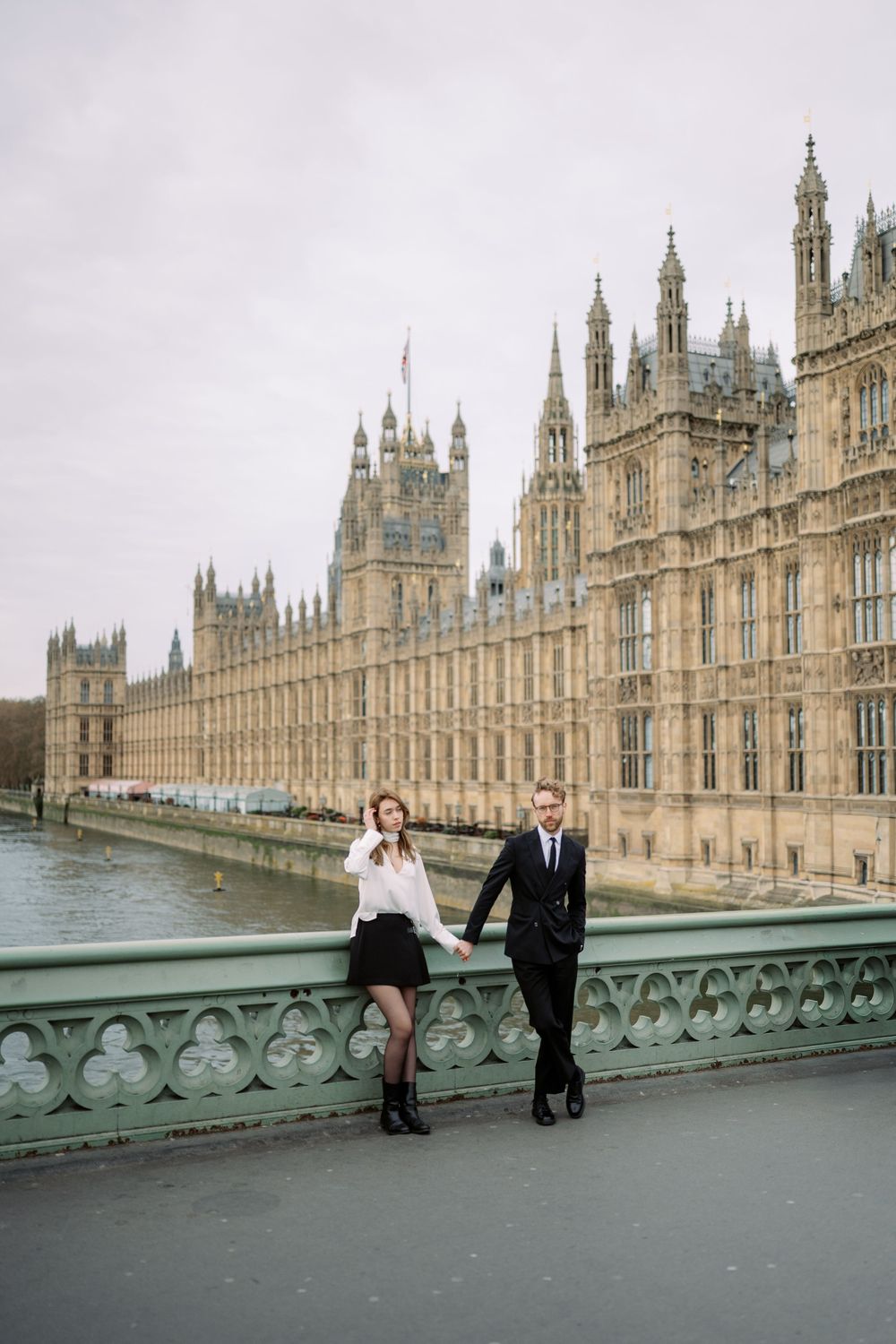 A couple poses on a bridge near the Houses of Parliament in London on an overcast day.