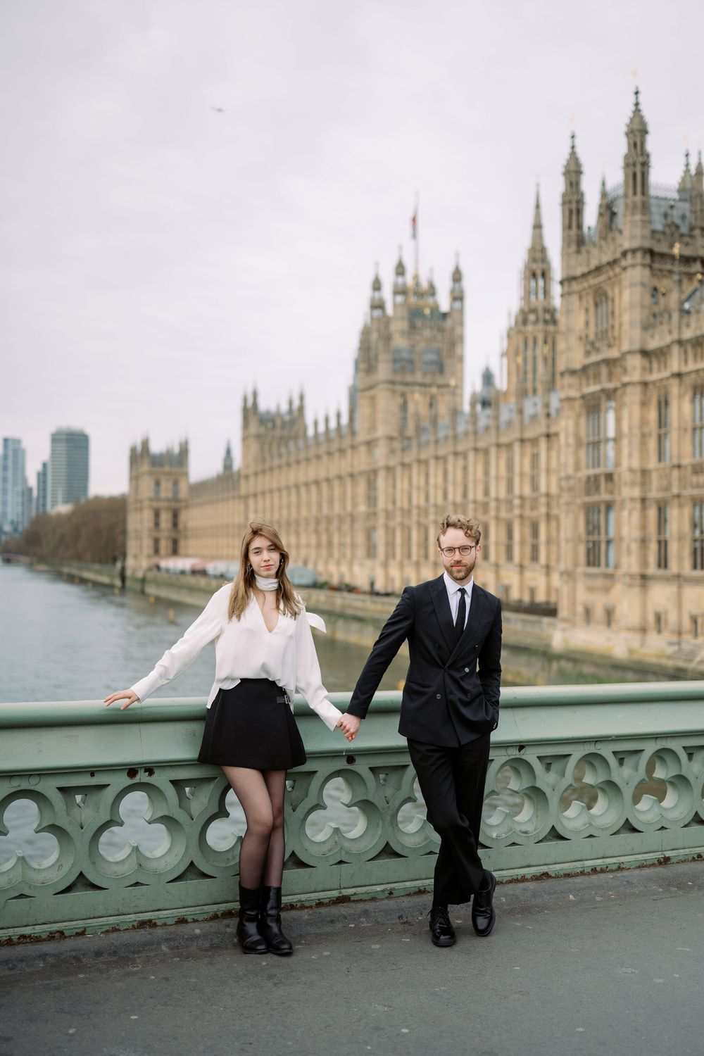 A couple poses on Westminster Bridge with the Houses of Parliament in the background on an overcast London day.