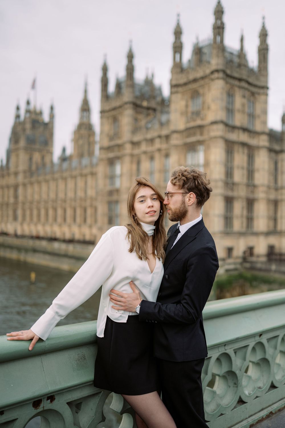 A romantic couple embraces on Westminster Bridge with the Houses of Parliament in the background on a cloudy day in London.
