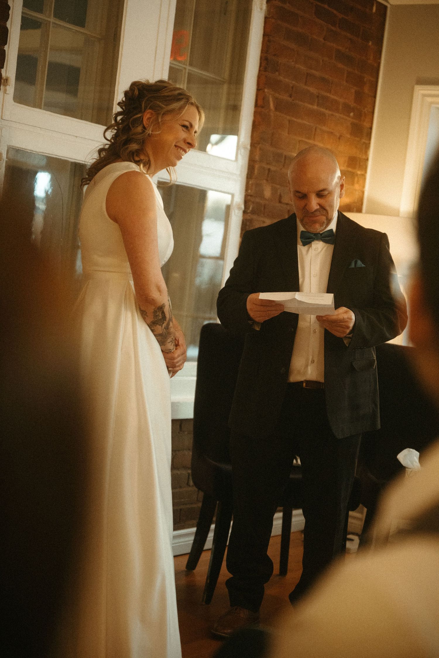 Bride in white gown smiles as groom in tuxedo reads vows during intimate wedding ceremony indoors.