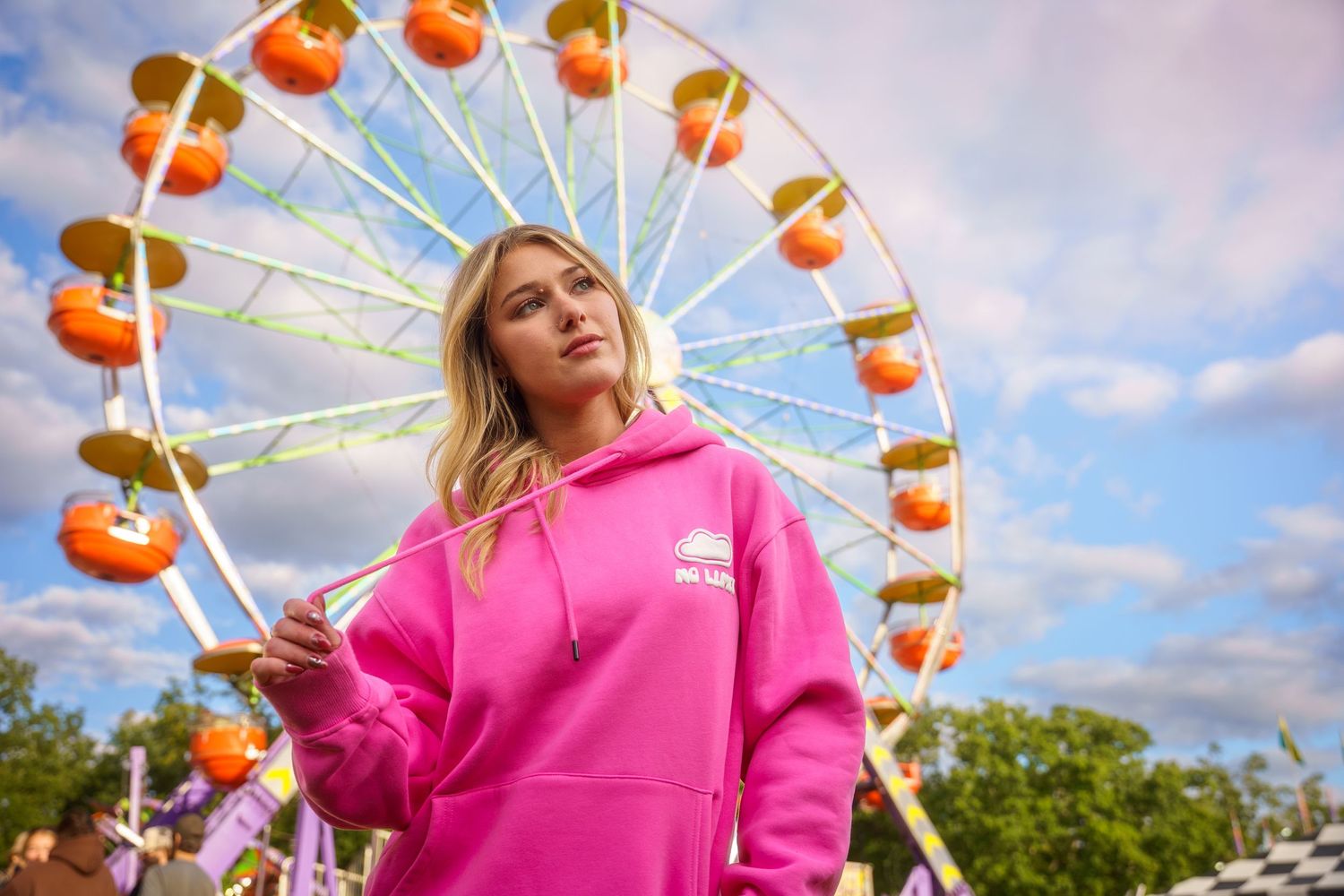 Blonde woman in bright pink hoodie standing in front of colorful carnival Ferris wheel outdoors.