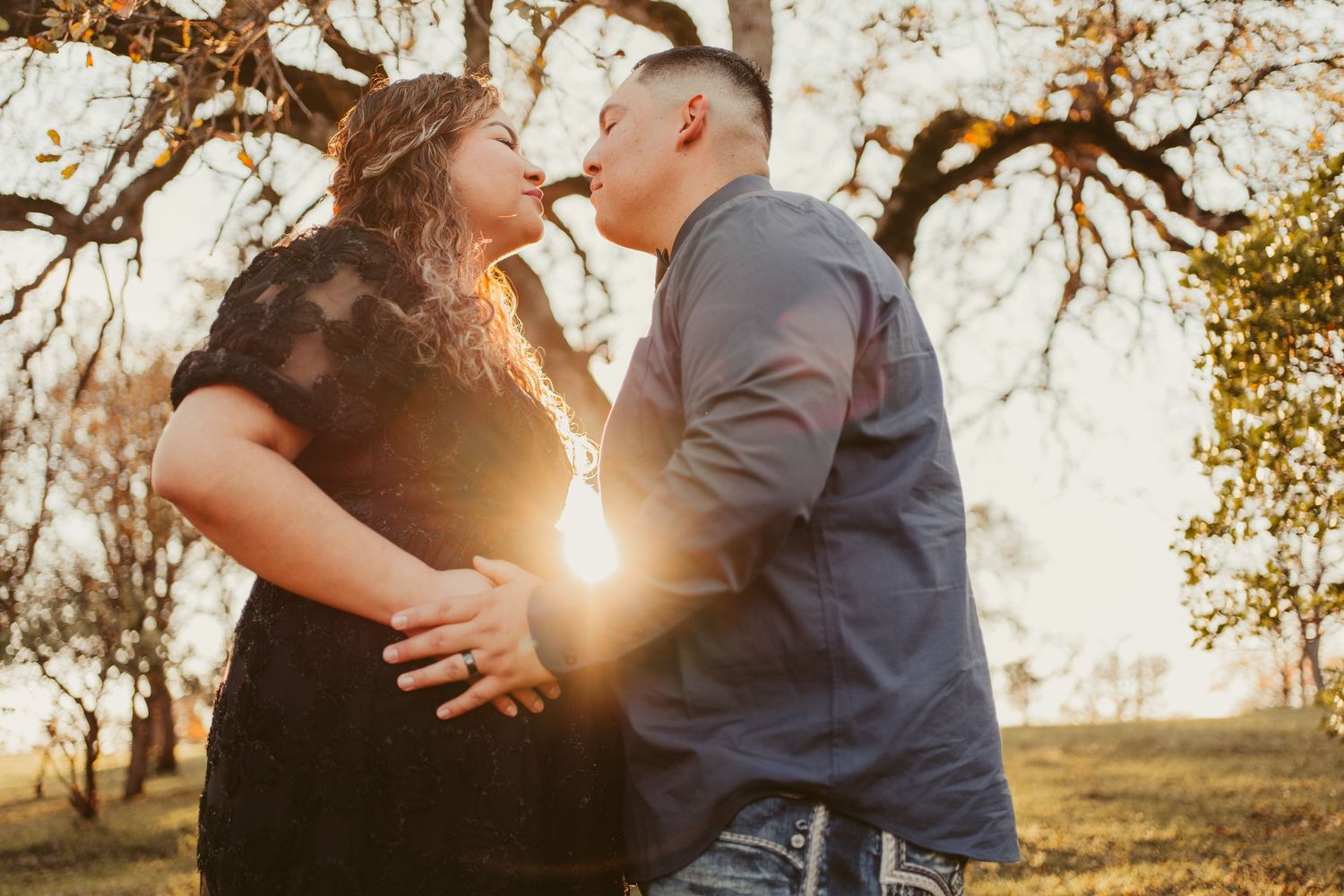 Couple sharing a romantic sunset kiss outdoors with golden backlight filtering through bare trees.