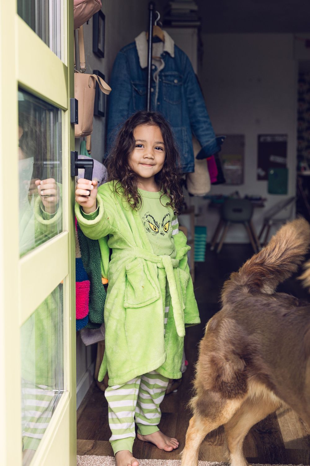 A young girl in a green Grinch robe opens a door while a dog stands nearby.