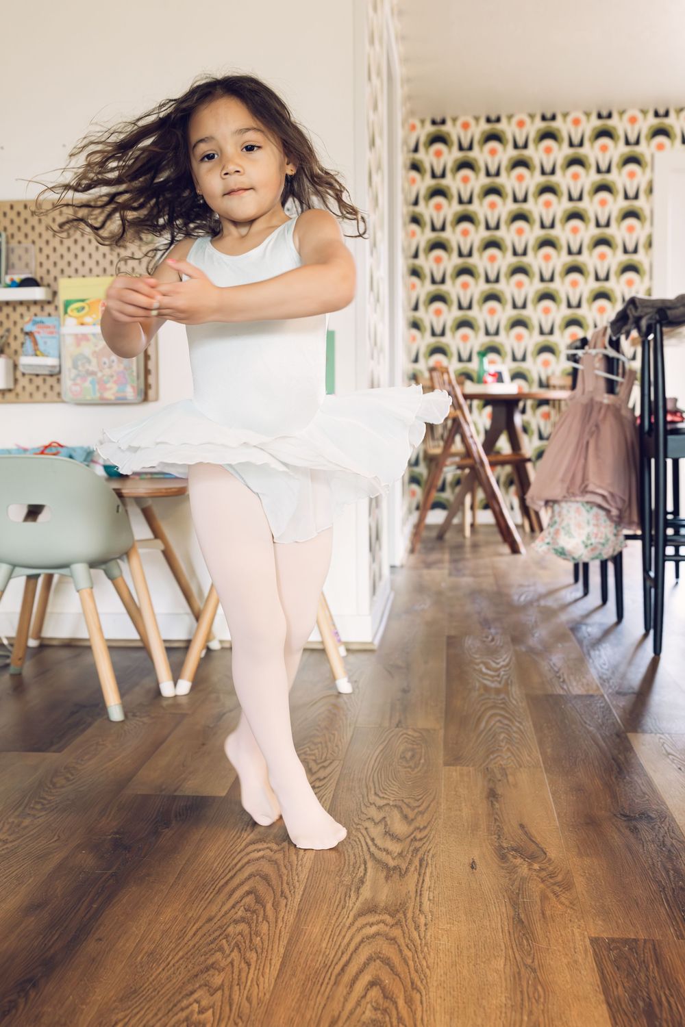 A young girl in a white ballet tutu twirls gracefully on a hardwood floor at home.