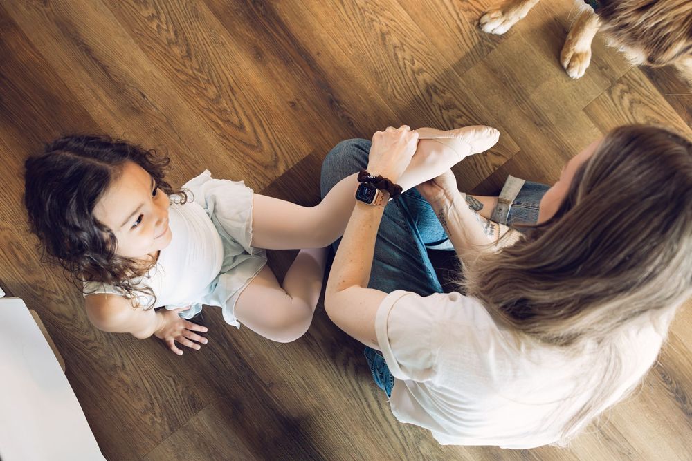 A mother and young daughter play together on a hardwood floor at home.