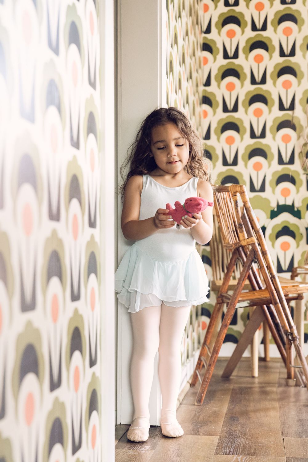 A young girl in a white ballet leotard holds a pink toy near a patterned wall.