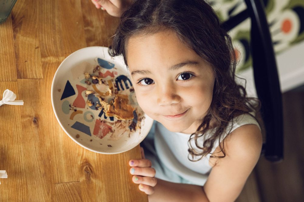 A smiling young girl holds up a colorful bowl of food at a wooden table.