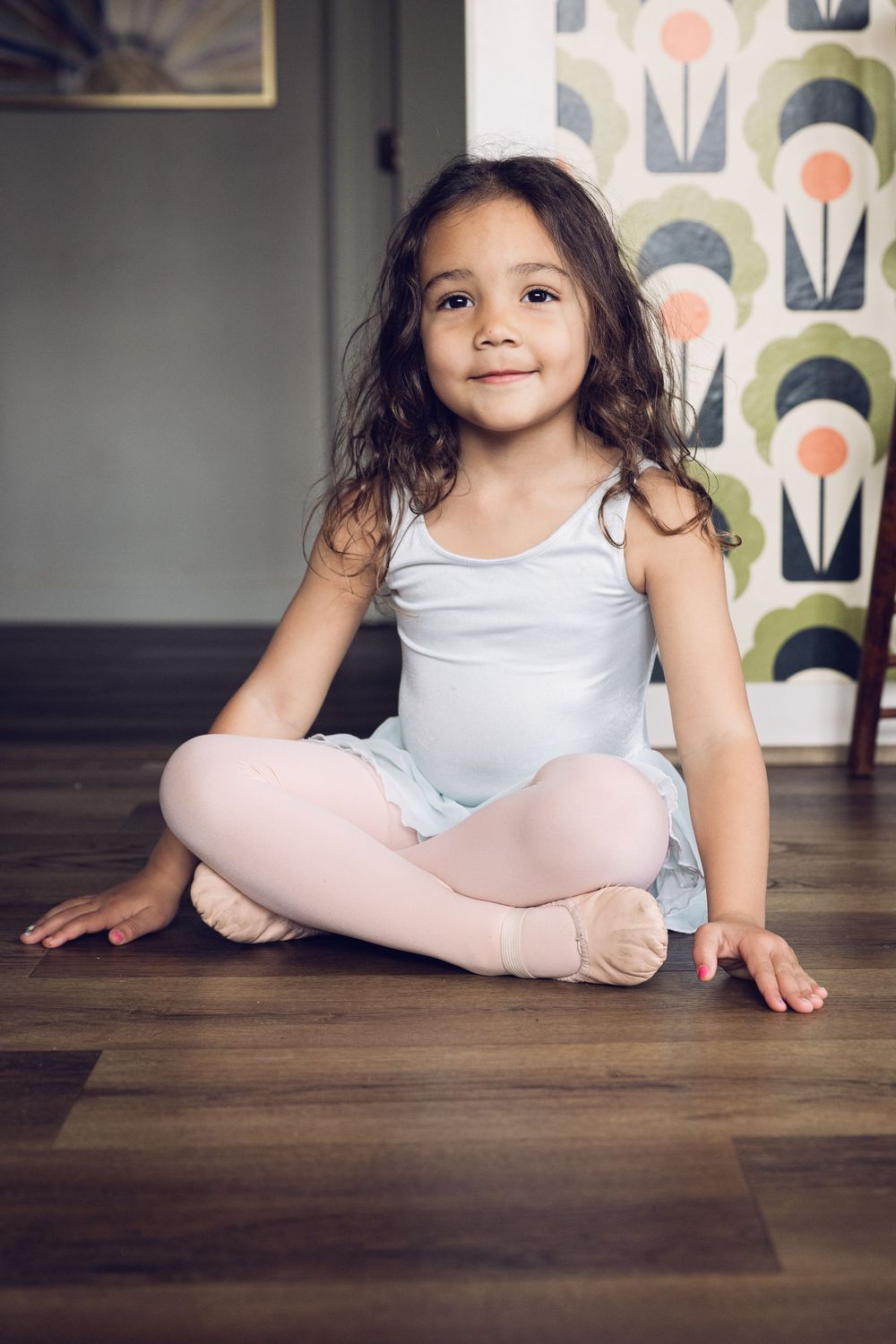 A young girl in a light blue ballet leotard sits cross-legged on a hardwood floor.