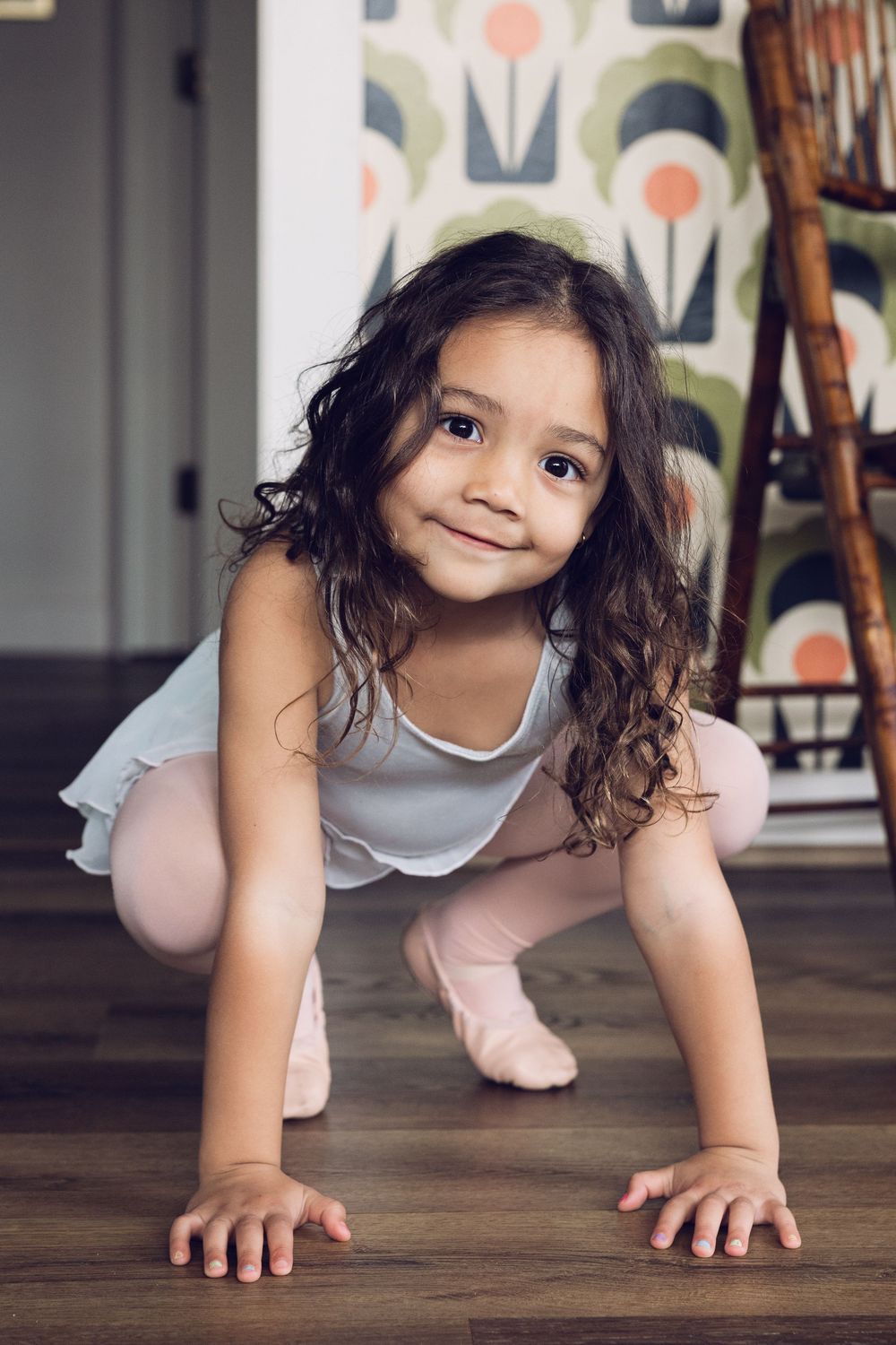 A smiling young girl with curly hair crouches on a hardwood floor wearing a white ballet leotard and pink tights.