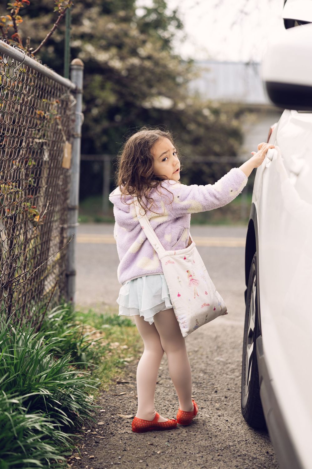 A little girl in a lavender jacket and tutu skirt touches a white car while carrying a floral bag.
