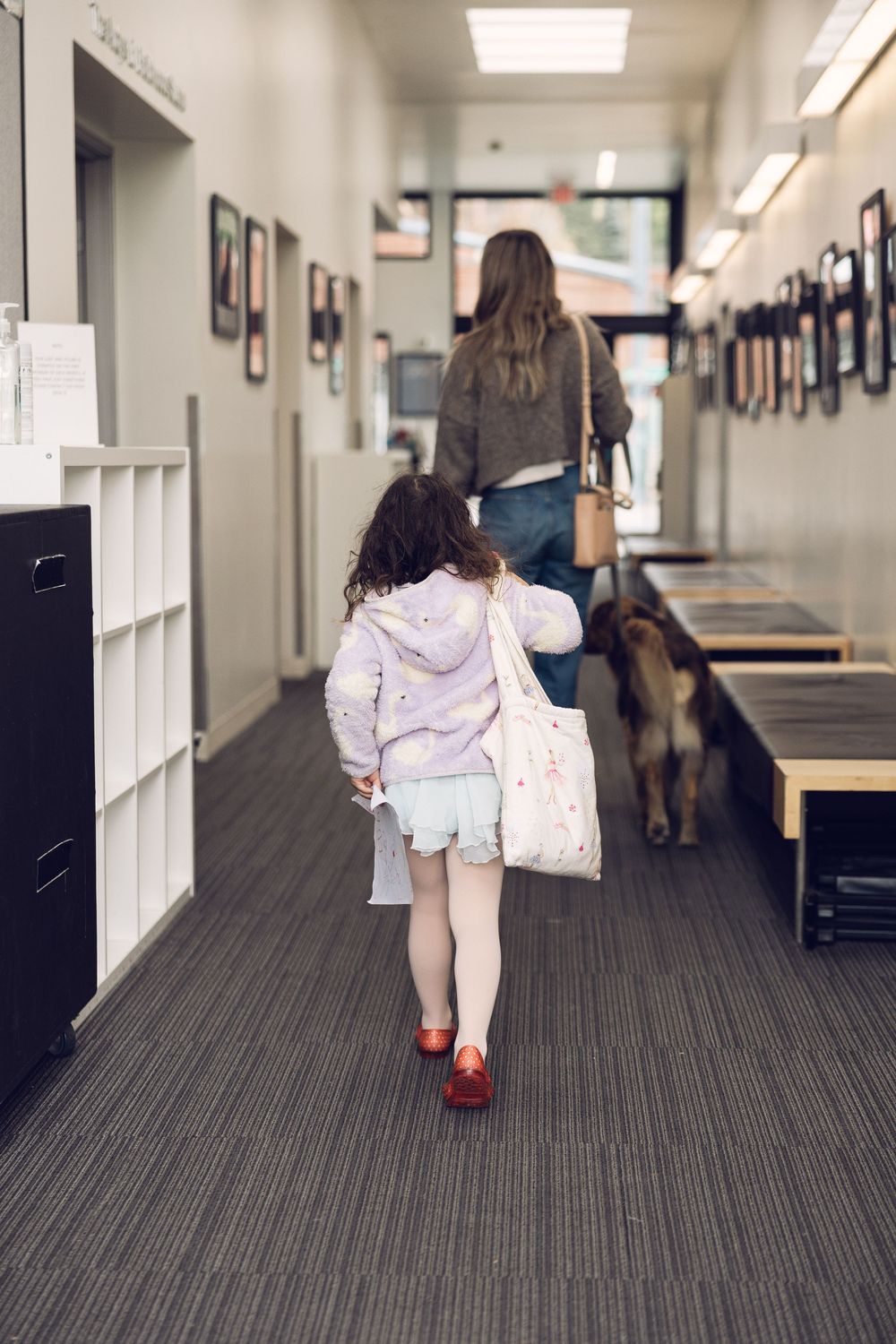 A young girl in a purple hoodie and tutu walks down a hallway with a woman and a dog ahead of her.