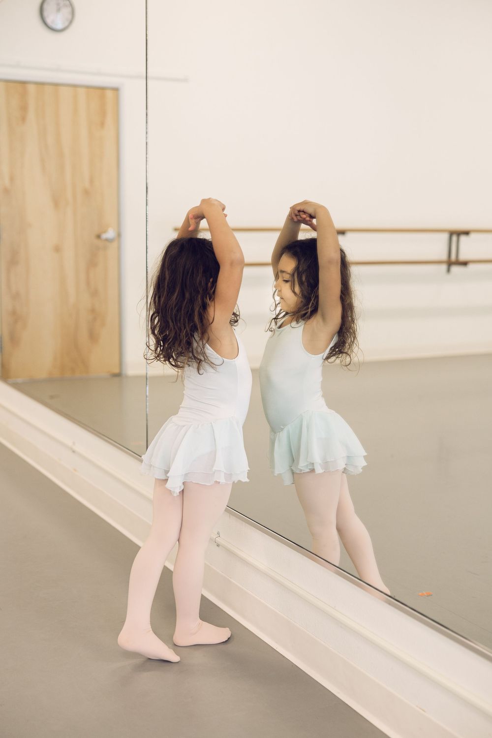 A young ballerina in a white tutu practices poses while looking at her reflection in a dance studio mirror.