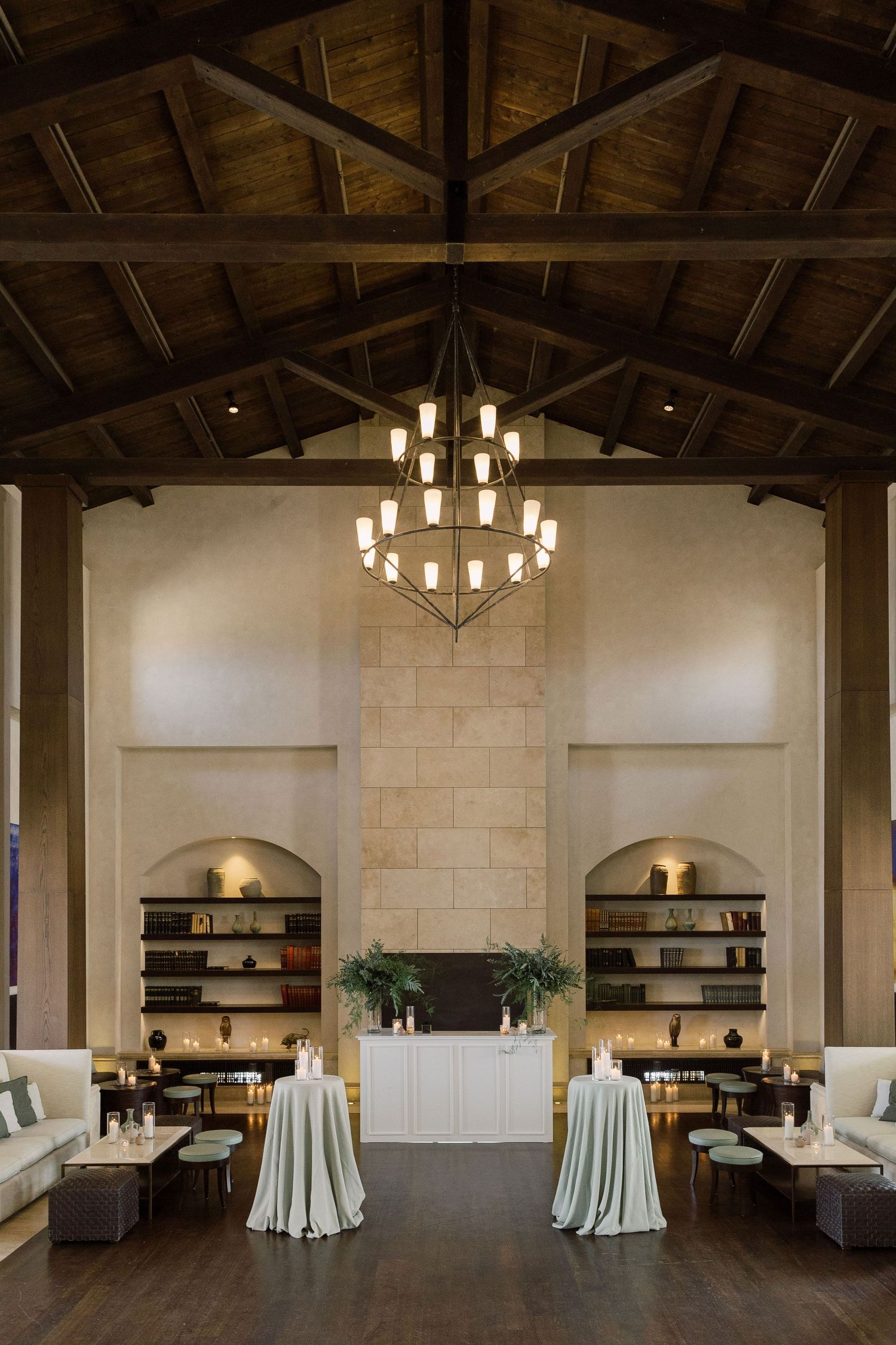 Modern rustic event venue interior with exposed wooden beams, chandeliers, and elegant table settings in neutral tones.