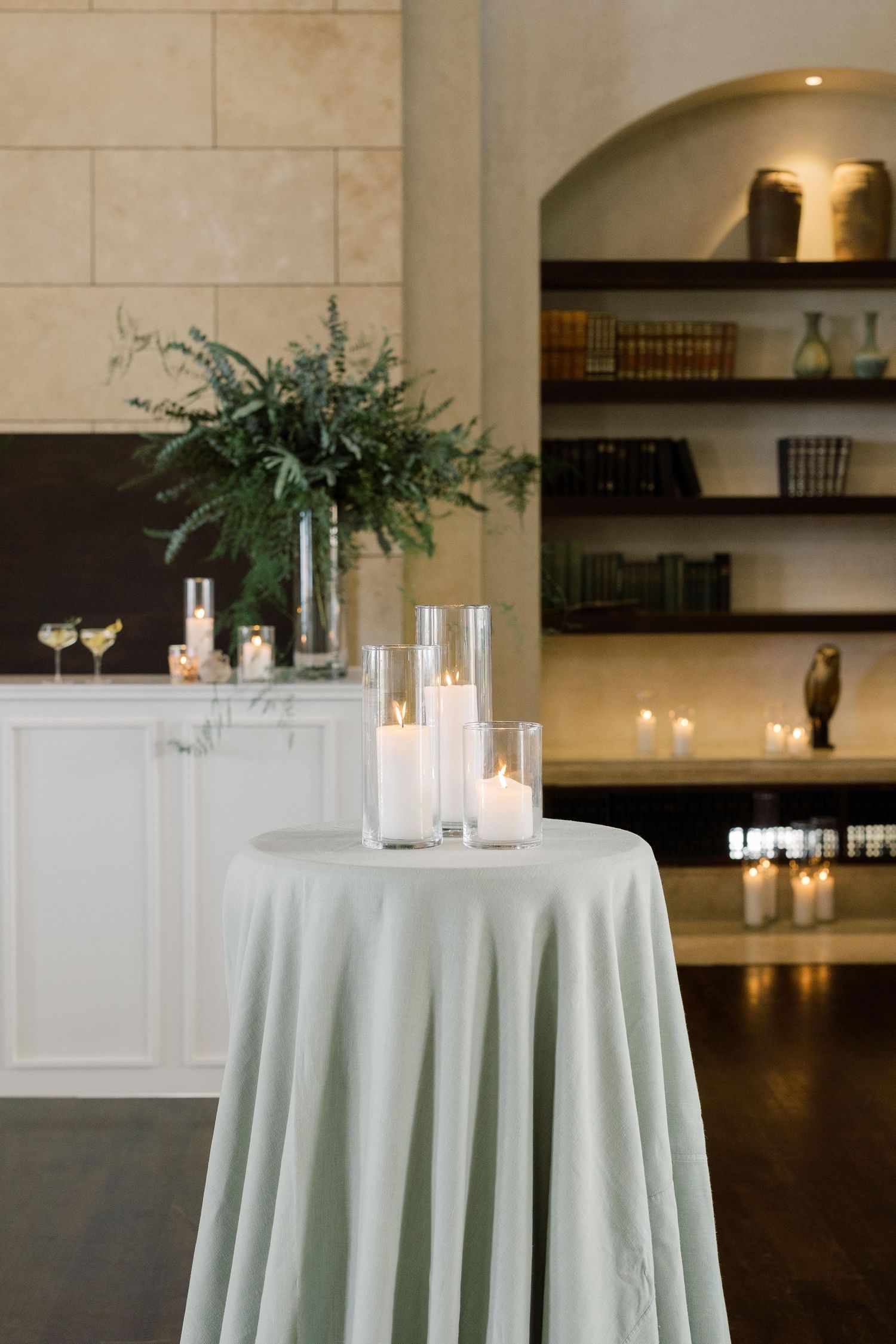 Elegant cocktail table with white tablecloth, glass candle holders and greenery arrangements in an upscale event space.