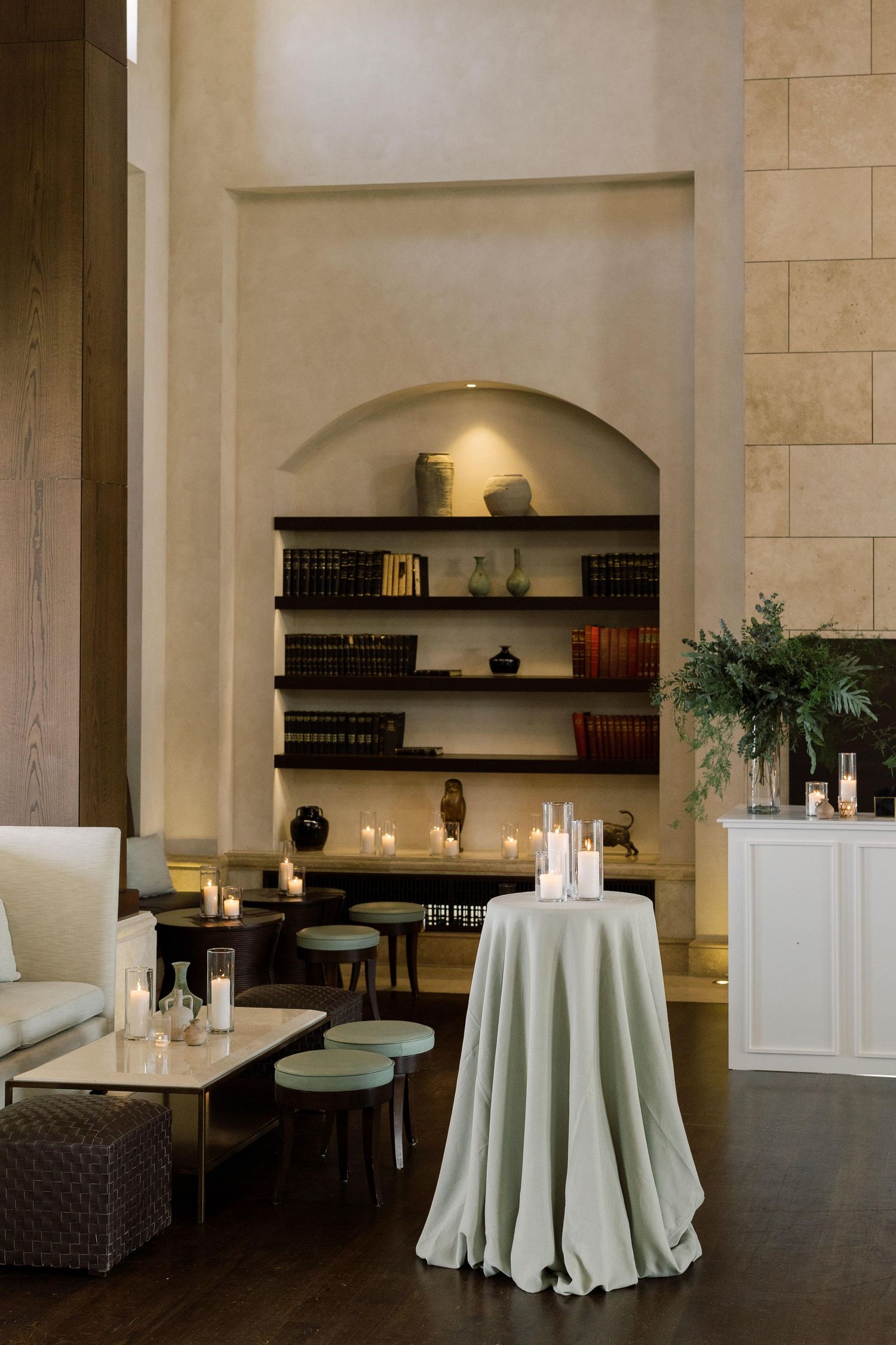 Modern event space with white round tables, dark wood shelving, and beige stone walls creating an elegant atmosphere.