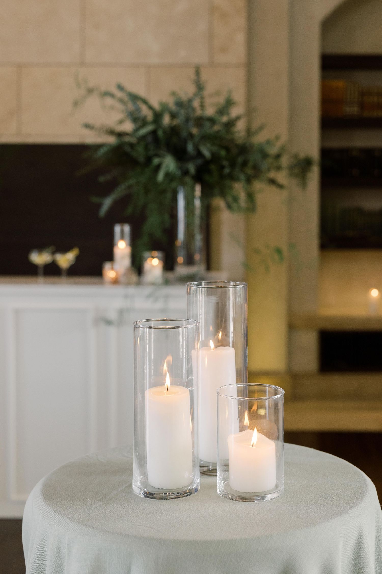 Lit white pillar candles in glass cylinders create an elegant ambiance on a white tablecloth.