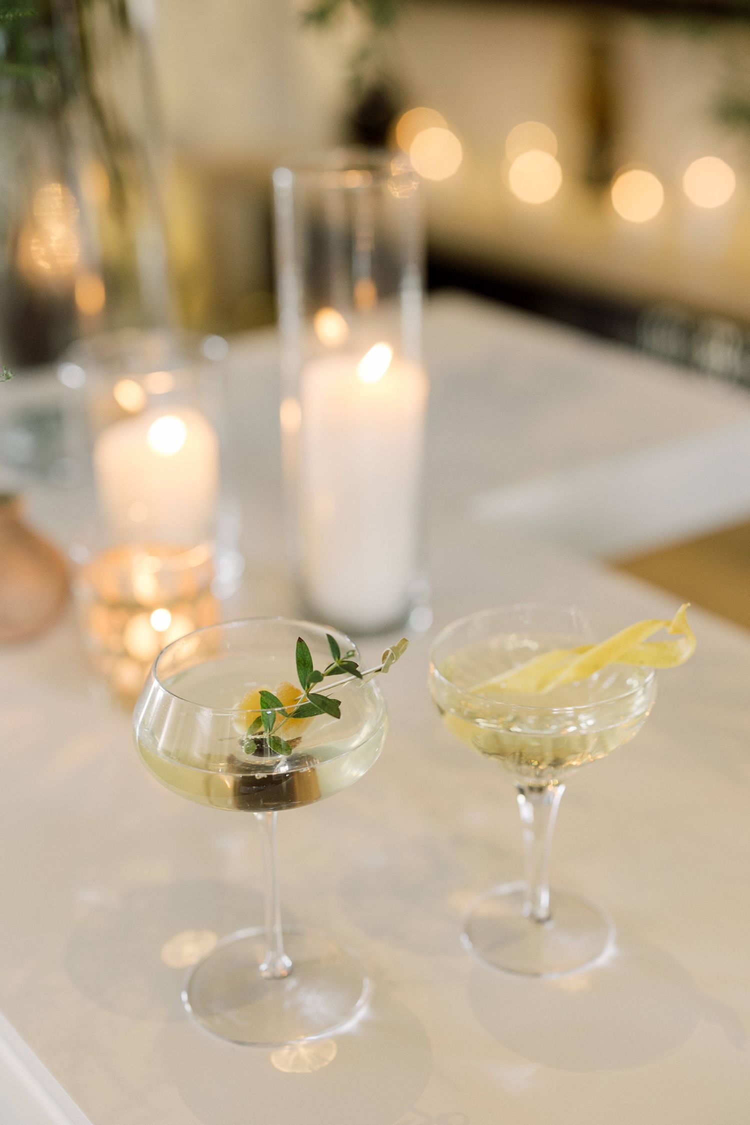 Elegant cocktail glasses garnished with citrus and herbs sit on a white tablecloth with soft glowing candlelight in the background.
