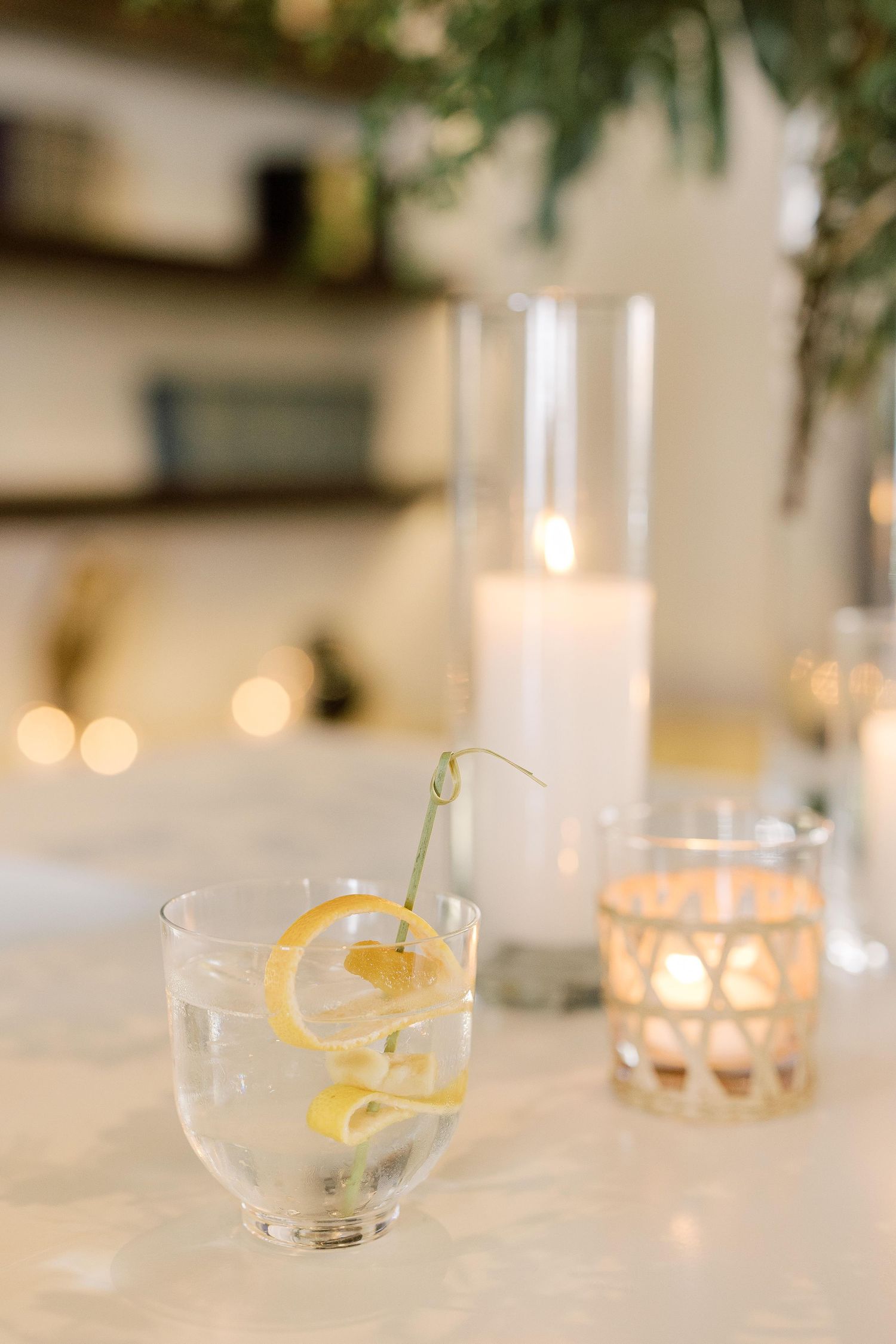 Row of coupe glasses with lemon twists and herbal garnishes arranged on an elegant white table with ambient lighting.