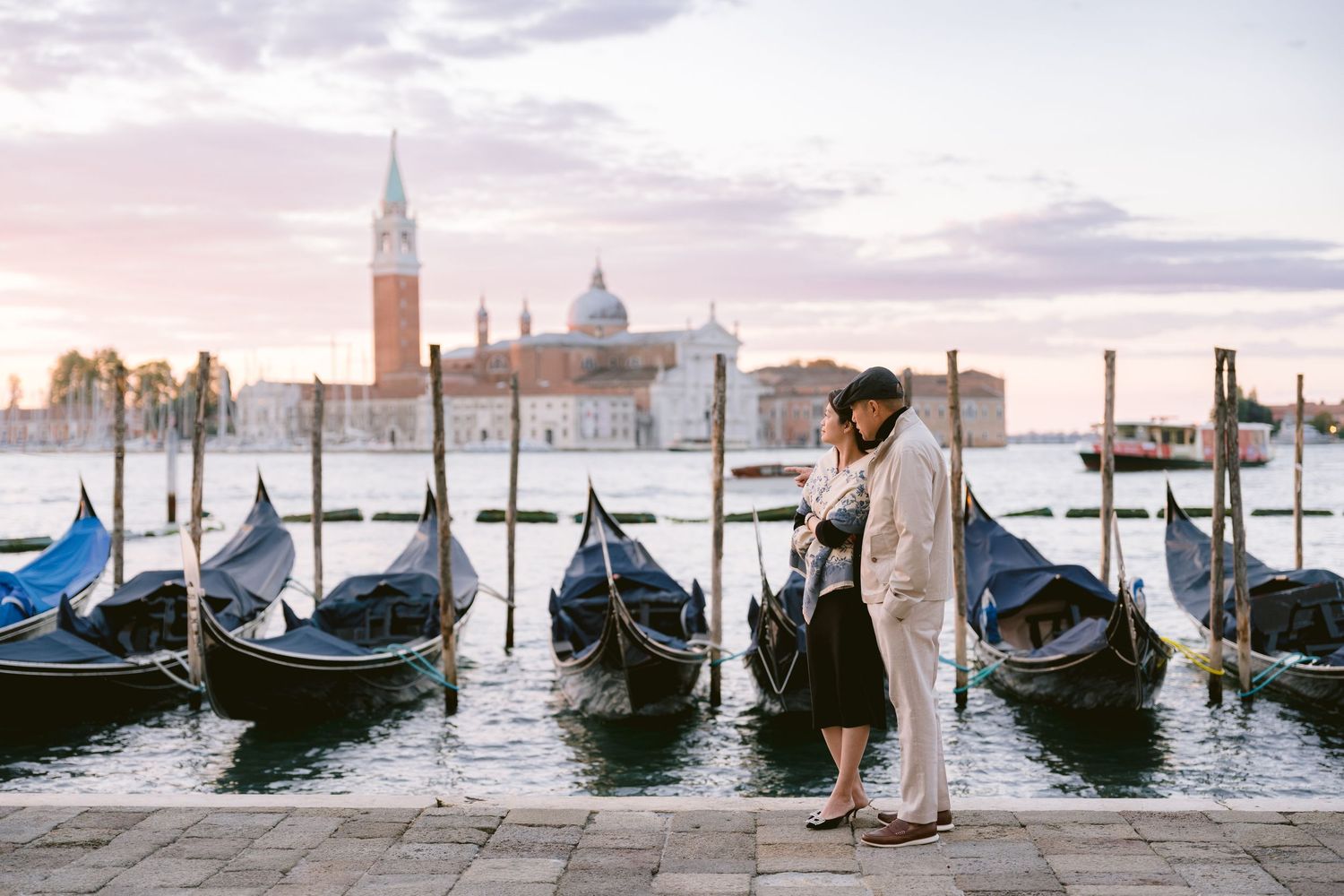 A series of black gondolas moored along the Venice waterfront with San Giorgio Maggiore church in the background at sunset.