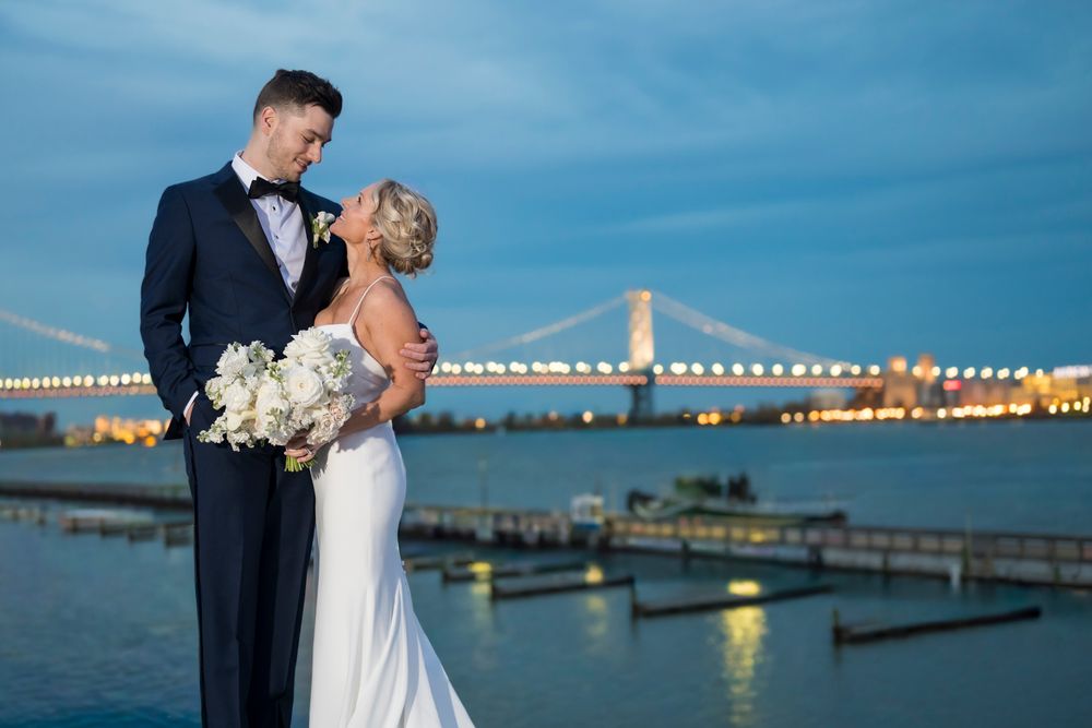 A couple shares an intimate moment at sunset by a waterfront marina with city lights twinkling in the background.