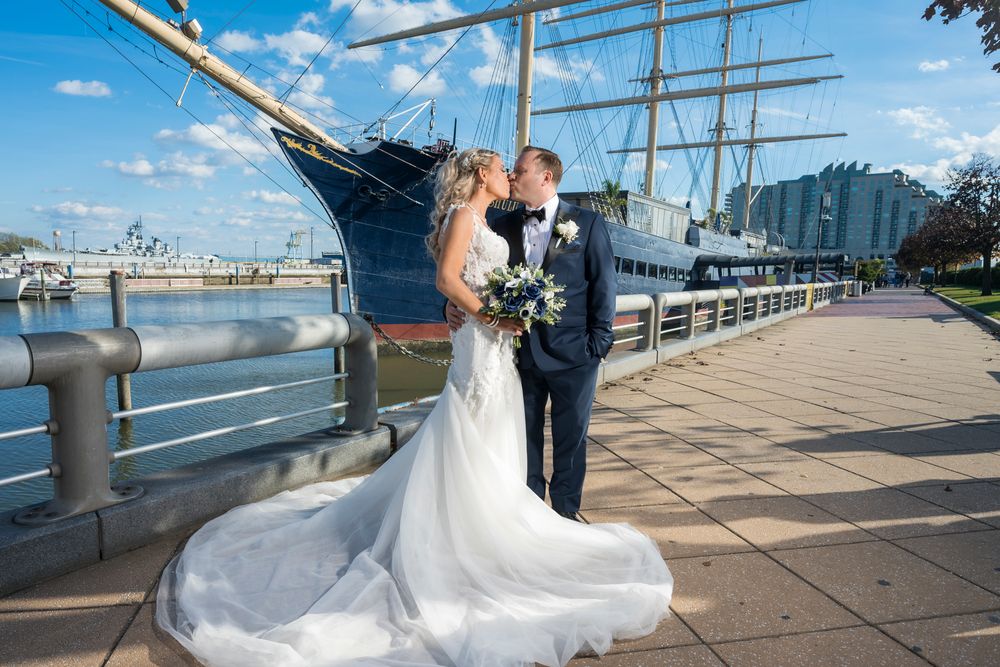 A wedding couple poses along a waterfront boardwalk with a tall ship and harbor in the background.