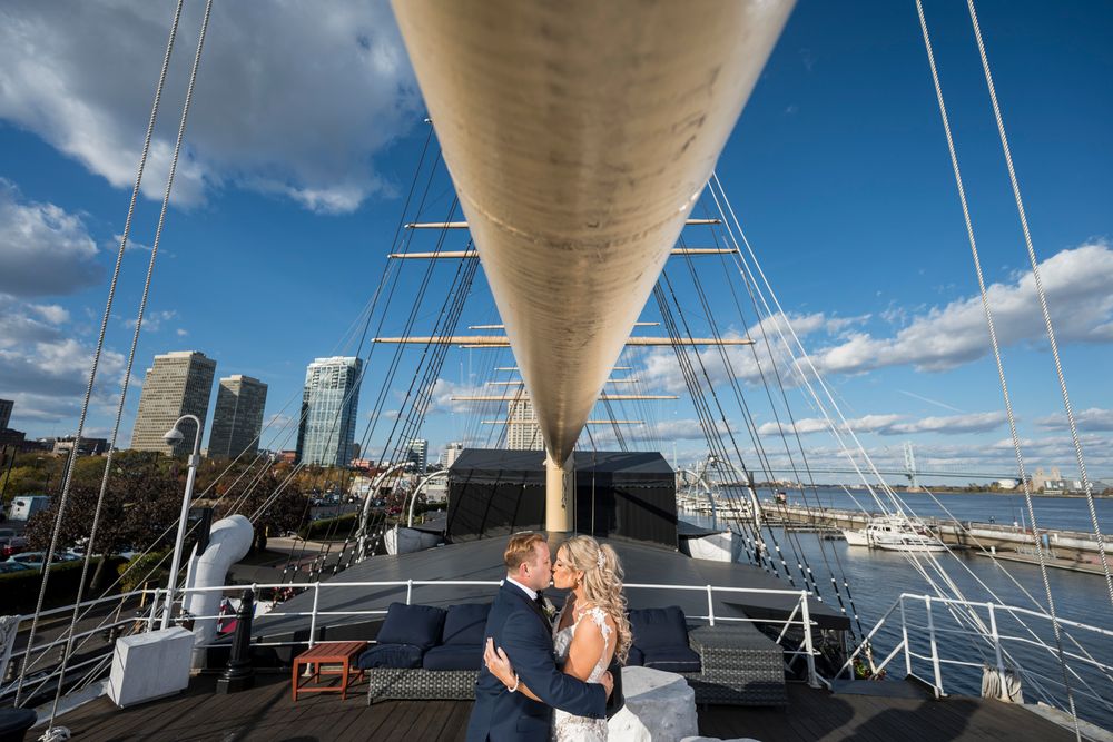 Two people embrace on the deck of a large sailing ship with tall masts and white sails against a blue sky.