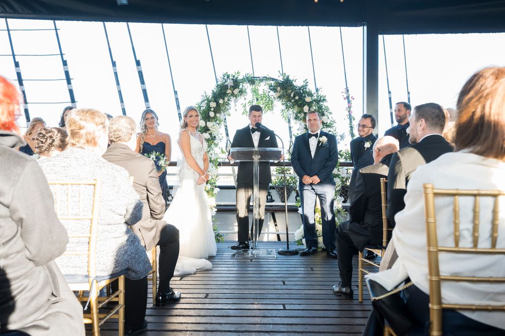 Wedding ceremony taking place on a covered deck with white floral arch and wooden chairs for guests.