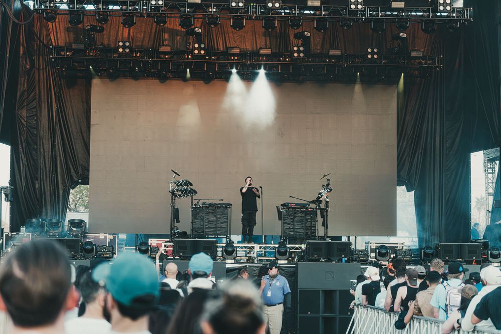 Concert performers on stage with spotlights and crowd at outdoor music festival venue during daytime performance.