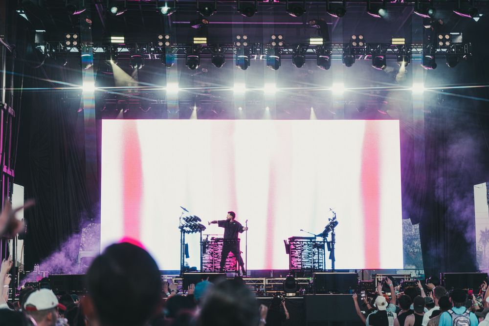 Concert stage setup with colorful lighting and video screens displaying pink and white stripes during a live performance.