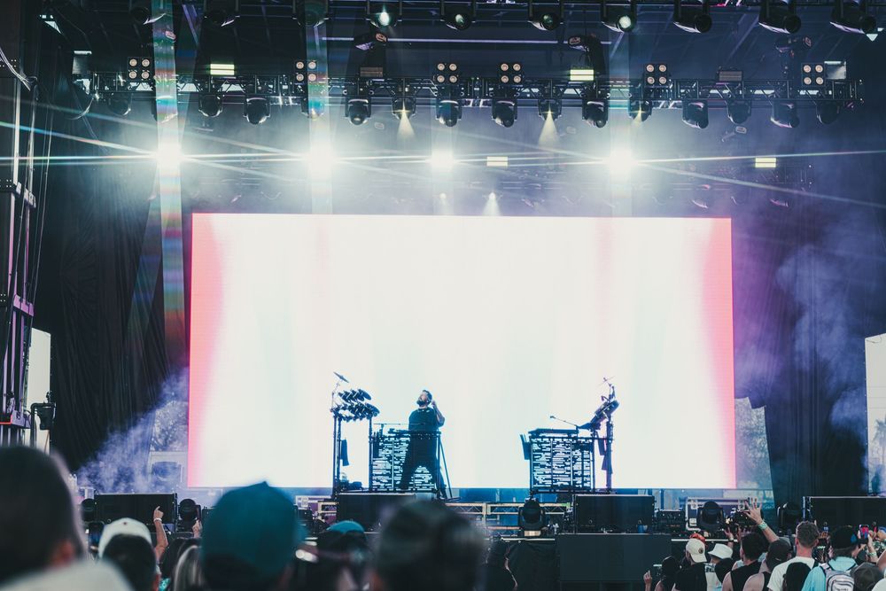 Wide concert venue with bright illuminated screens showing vertical stripes and atmospheric smoke effects.