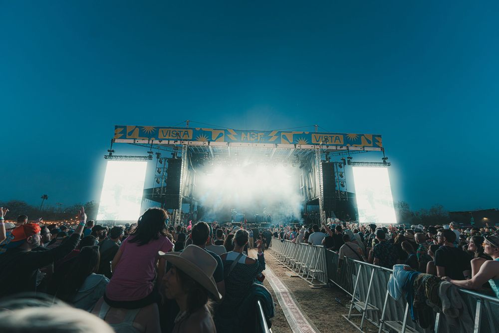 A large crowd gathers at an outdoor concert venue with stage lights illuminating the scene at dusk.