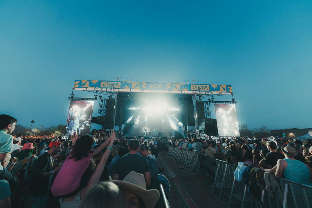 Outdoor music festival crowd gathered in front of illuminated stage with bright lights under blue evening sky.