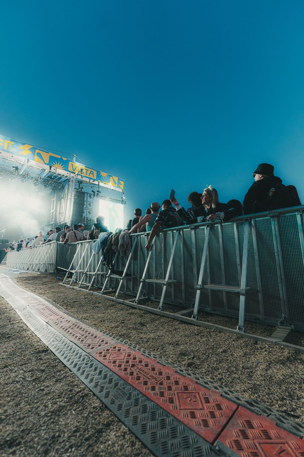 Metal crowd control barriers with red anti-slip panels line up against a dark sky with bright lights in the background.