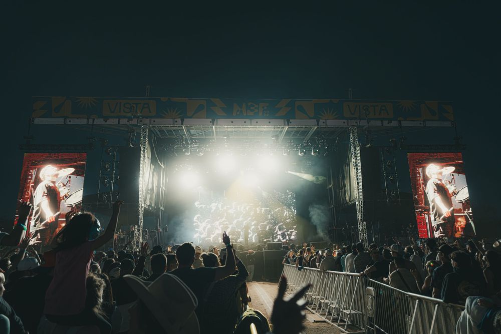 Concert crowd silhouettes gather in front of illuminated stage with dramatic lighting and fog effects during live performance.
