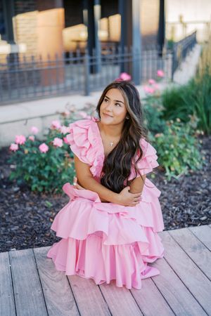A pink ruffled dress is worn while sitting on wooden decking with pink flowers in the background.