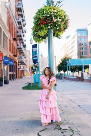 A pink ruffled dress stands out against urban architecture and a hanging flower basket on a city street.