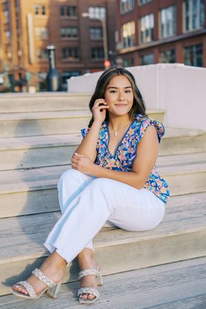 Casual portrait on wooden steps wearing white pants and floral top in urban setting.